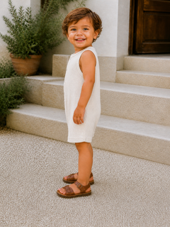 Smiling child in a light outfit stands on steps with greenery in the background, wearing brown sandals.