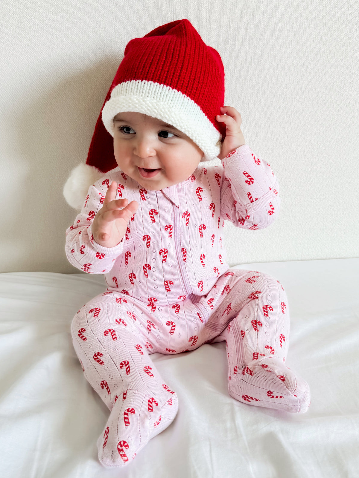 Infant wearing a festive red and white Santa hat and pink candy cane pajamas, sitting on a white blanket.