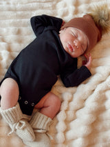 Newborn baby wearing a black onesie and brown hat, peacefully sleeping on a soft blanket.