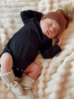 Newborn baby wearing a black onesie and brown hat, peacefully sleeping on a soft blanket.