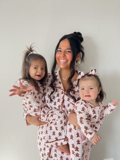 Woman holding two smiling toddlers, all in matching pink pajamas with a gingerbread design against a neutral backdrop.
