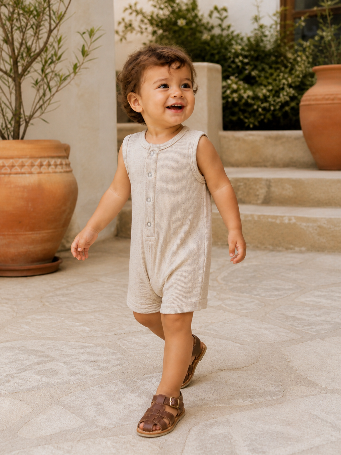 Smiling child in a beige romper and brown sandals walking on a stone pathway near a plant pot.