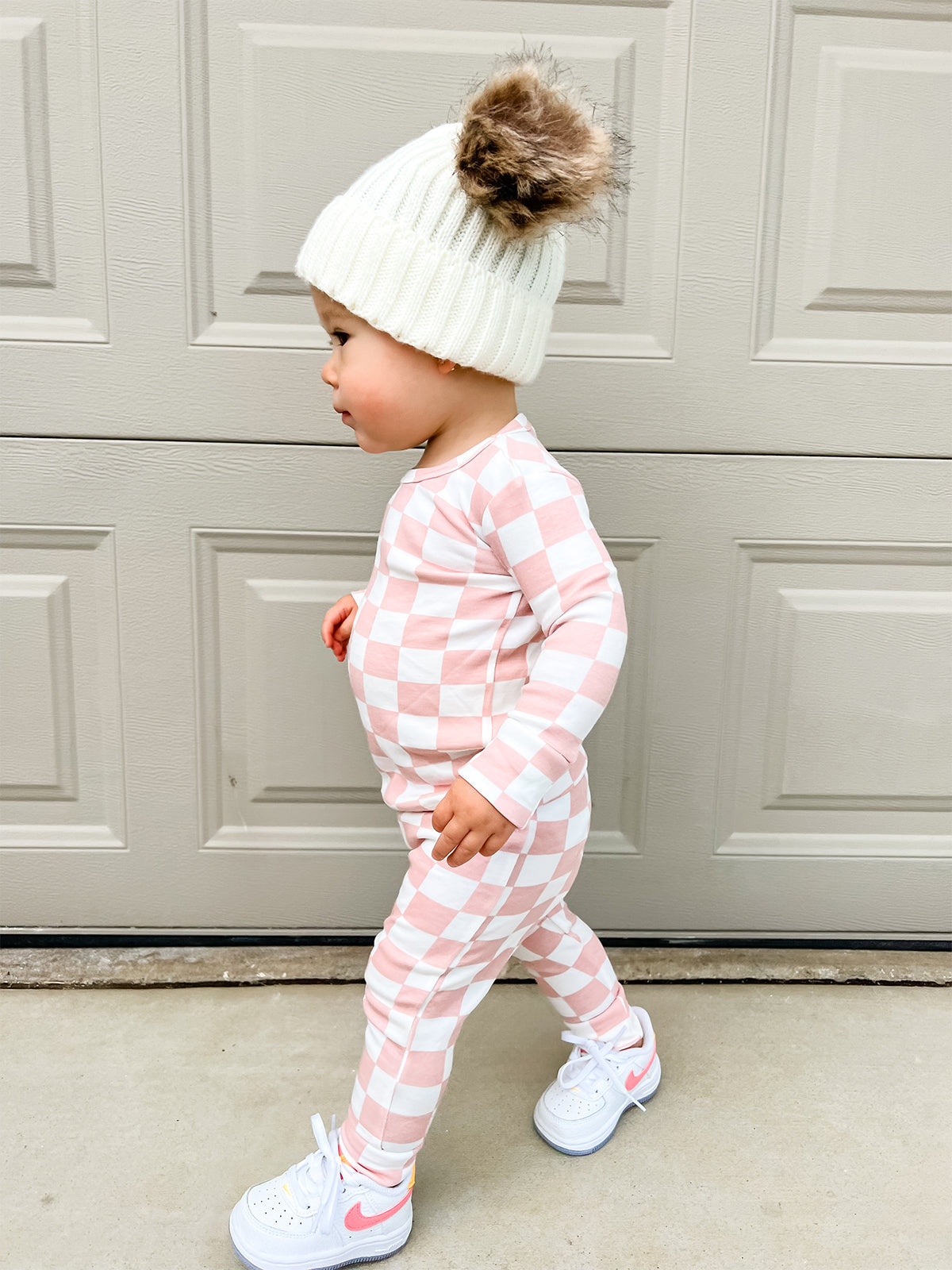 Toddler in pink checkered outfit and white beanie with pom-pom, walking beside a garage door.
