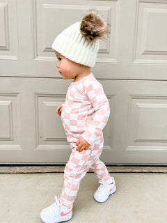Toddler in pink checkered outfit and white beanie with pom-pom, walking beside a garage door.
