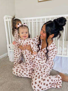 Mother and baby wearing matching pink gingerbread-themed pajamas, seated near a crib, with a playful atmosphere.