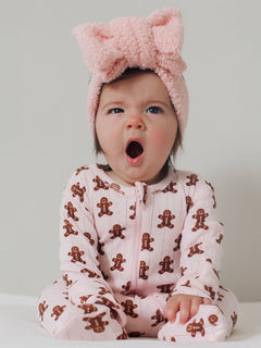 Baby in a pink gingerbread onesie yawns, wearing a fluffy pink headband, sitting against a neutral background.