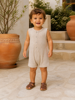 Smiling child in a beige romper stands on stone steps, with greenery and terracotta pots in the background.