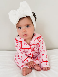 Baby wearing pink candy cane pajamas and a white bow headband, sitting on a bed with a neutral background.
