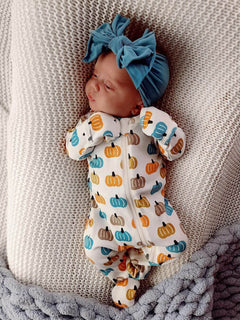 Baby dressed in a pumpkin-patterned onesie, wearing a teal bow headband, resting on a cozy blanket.