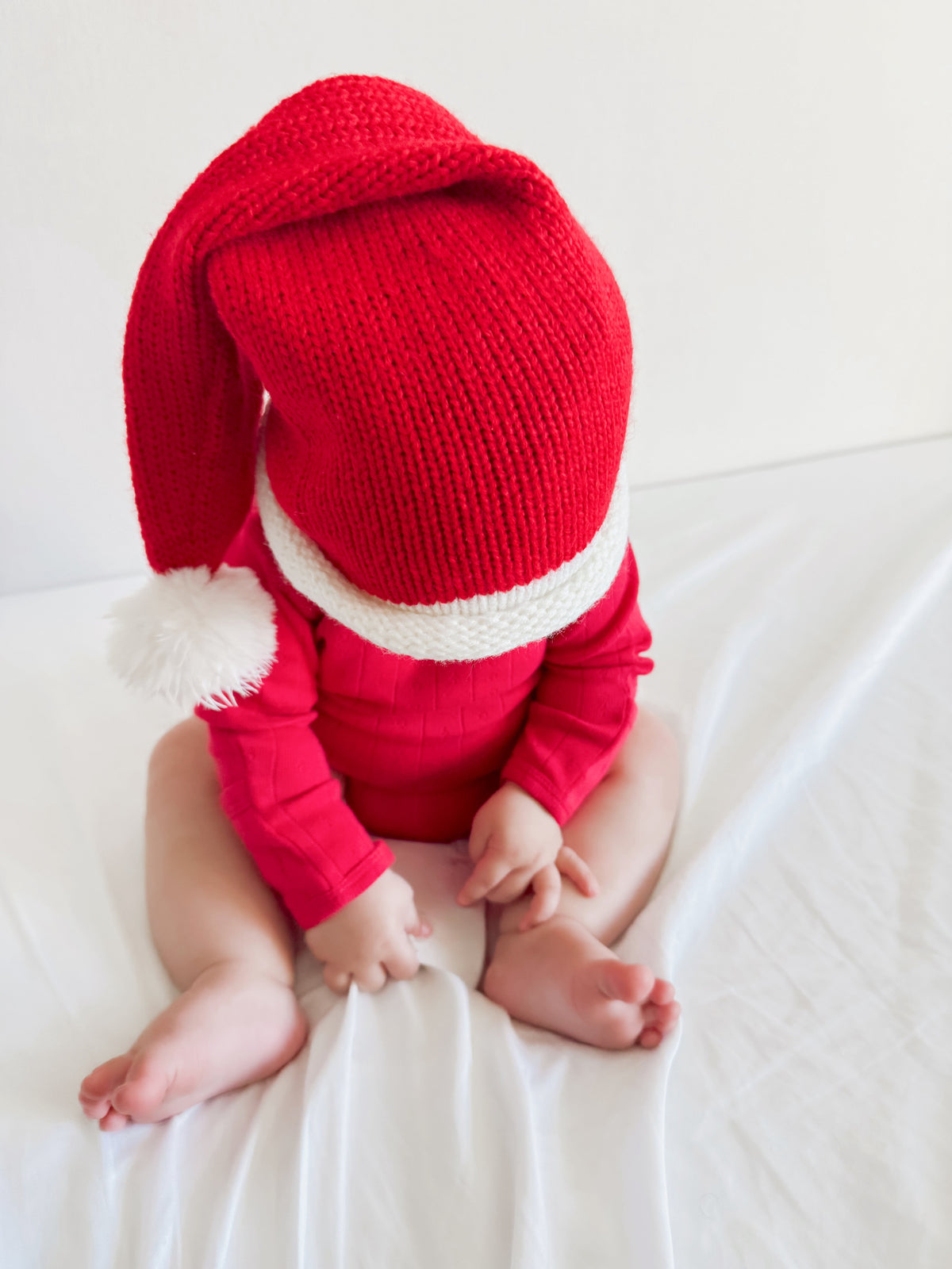 Baby wearing a red Santa hat and sweater, sitting on a white blanket with bare feet.