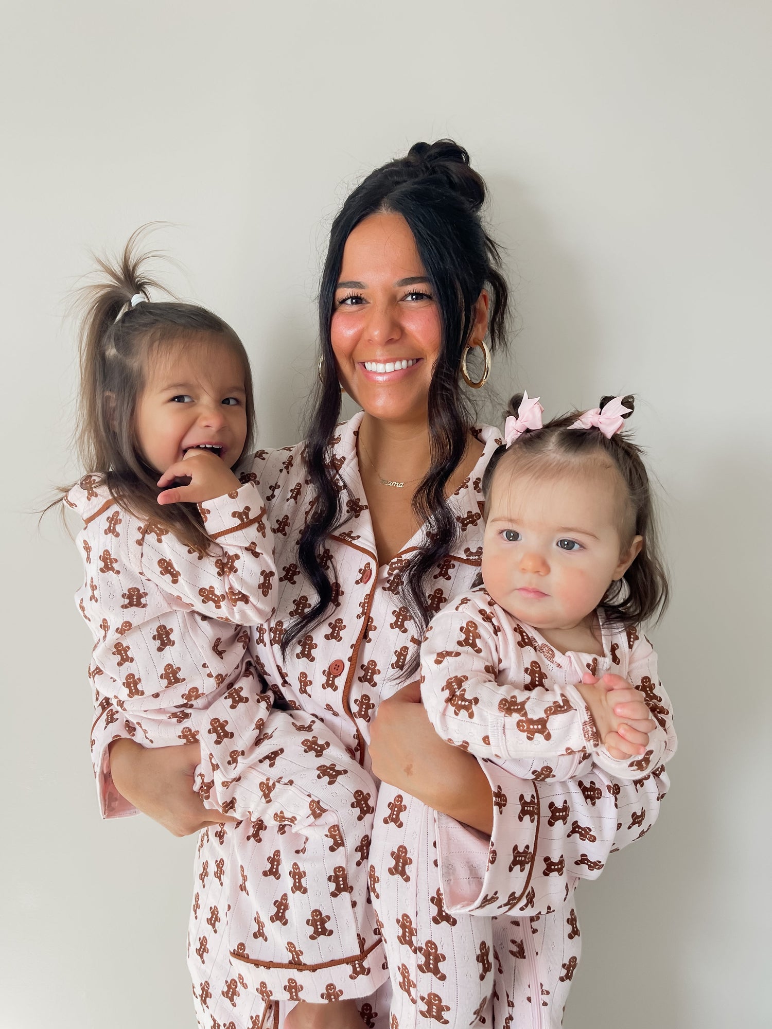 Smiling woman holds two young girls in matching gingerbread-themed pajamas against a neutral background.