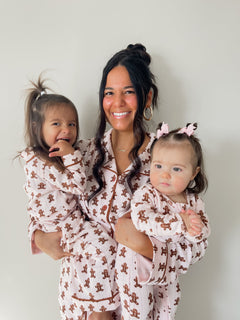 Smiling woman holds two young girls in matching gingerbread-themed pajamas against a neutral background.