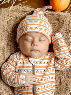Sleeping baby in pumpkin-themed outfit on a burlap backdrop, with a pumpkin nearby.