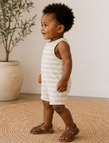 Smiling toddler in a striped romper and sandals, standing by a potted plant on a woven rug.