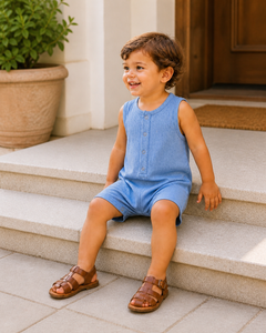 Smiling child in blue romper sitting on steps, wearing brown sandals with greenery and a door in the background.