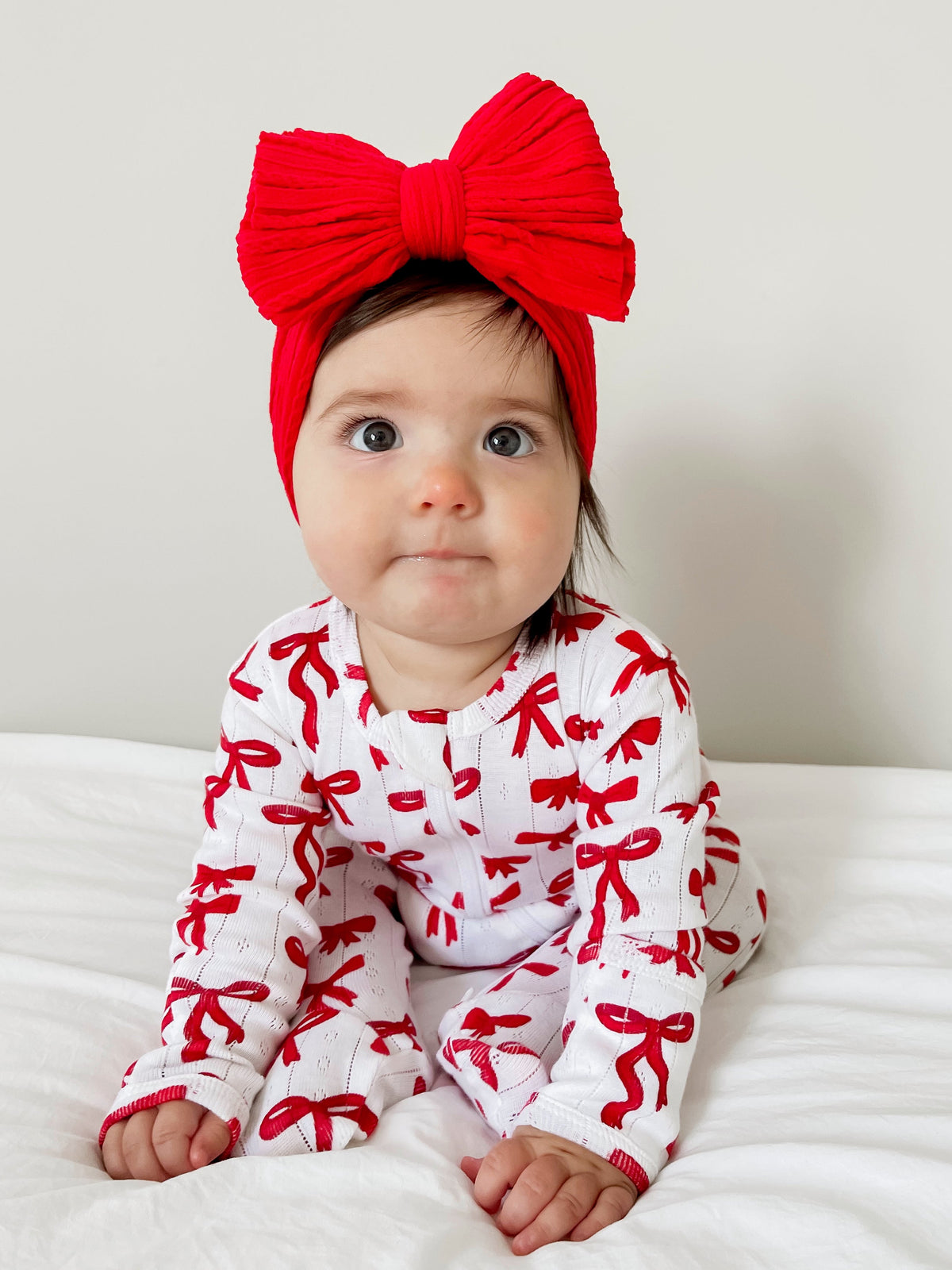 Baby wearing white outfit with red bows and a large red bow headband, sitting on a white bed.