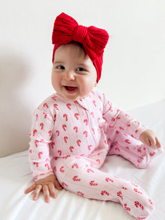 Smiling baby in a pink candy-cane outfit with a large red bow, sitting on a white bed.