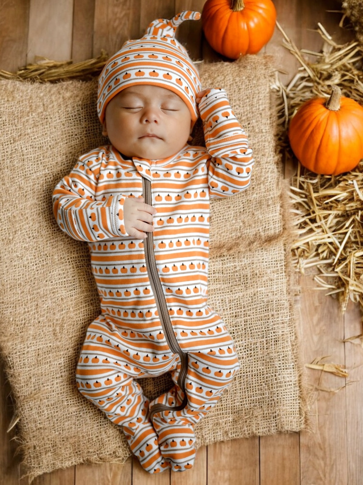 Sleeping baby in pumpkin-patterned outfit, surrounded by pumpkins on a straw mat. Warm autumn theme.