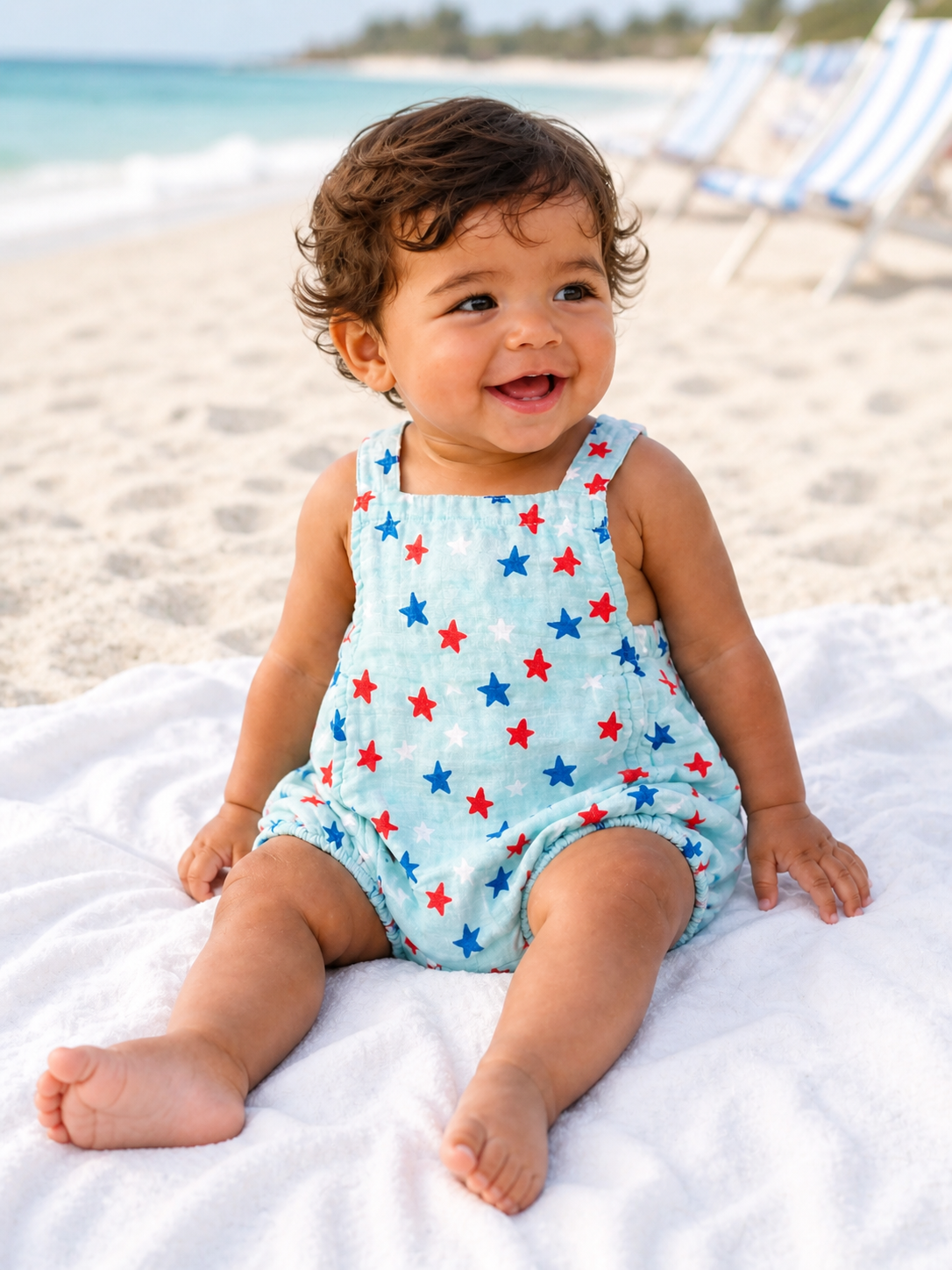 Smiling baby in a star-patterned romper seated on a beach towel, with a sandy beach and ocean in the background.