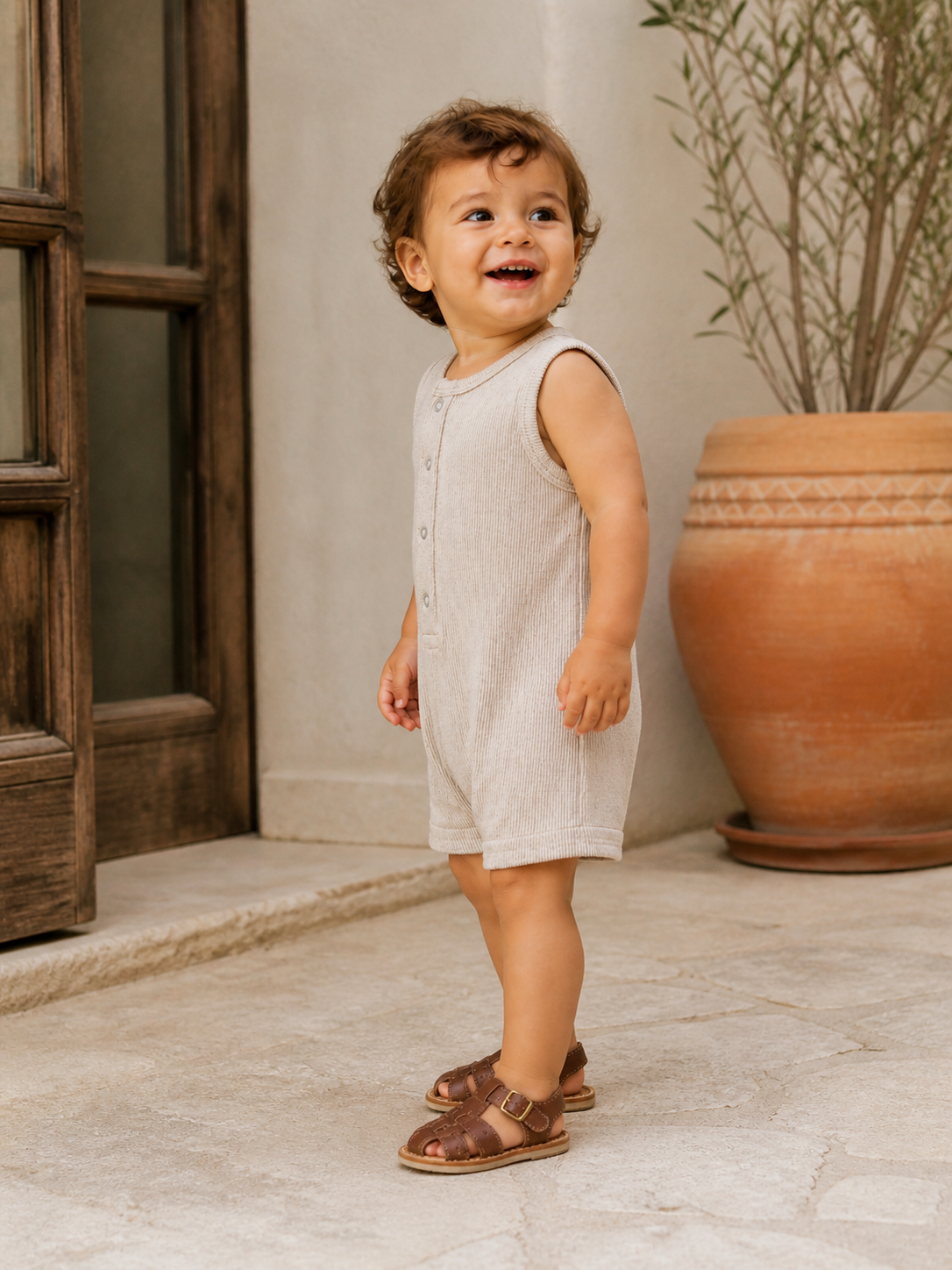 Smiling toddler in a beige romper stands on stone steps by a terracotta pot, wearing brown sandals.