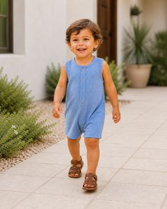 Smiling child in a blue romper walking on a sunny path, surrounded by greenery and modern architecture.