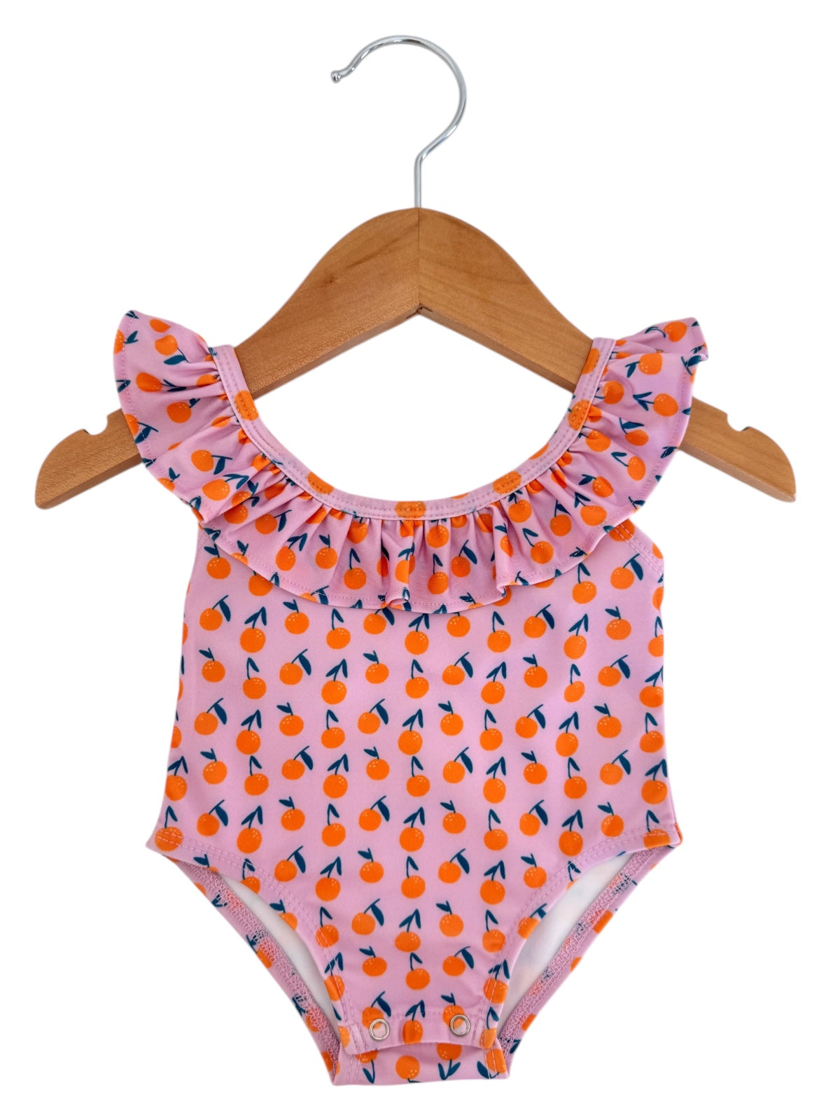 Pink swimsuit with orange cherry print and ruffled neckline, hanging on a wooden hanger against a white background.