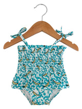 Blue swimsuit with banana print, smocked bodice, and shoulder ties, hanging on a wooden hanger.