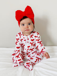 Infant girl in a white outfit with red bows, wearing a large red bow headband, seated on a white bed.