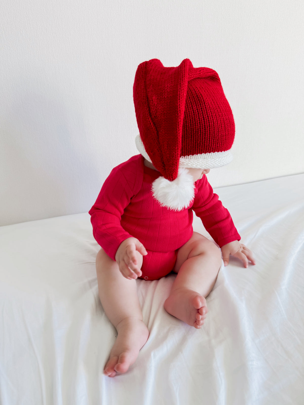 Baby wearing a red holiday outfit and oversized Santa hat, seated on a bed with white sheets.