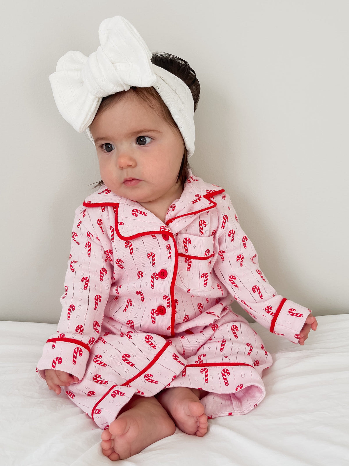 Toddler in pink candy-cane pajamas and a white headband, sitting on a white bed with a neutral background.