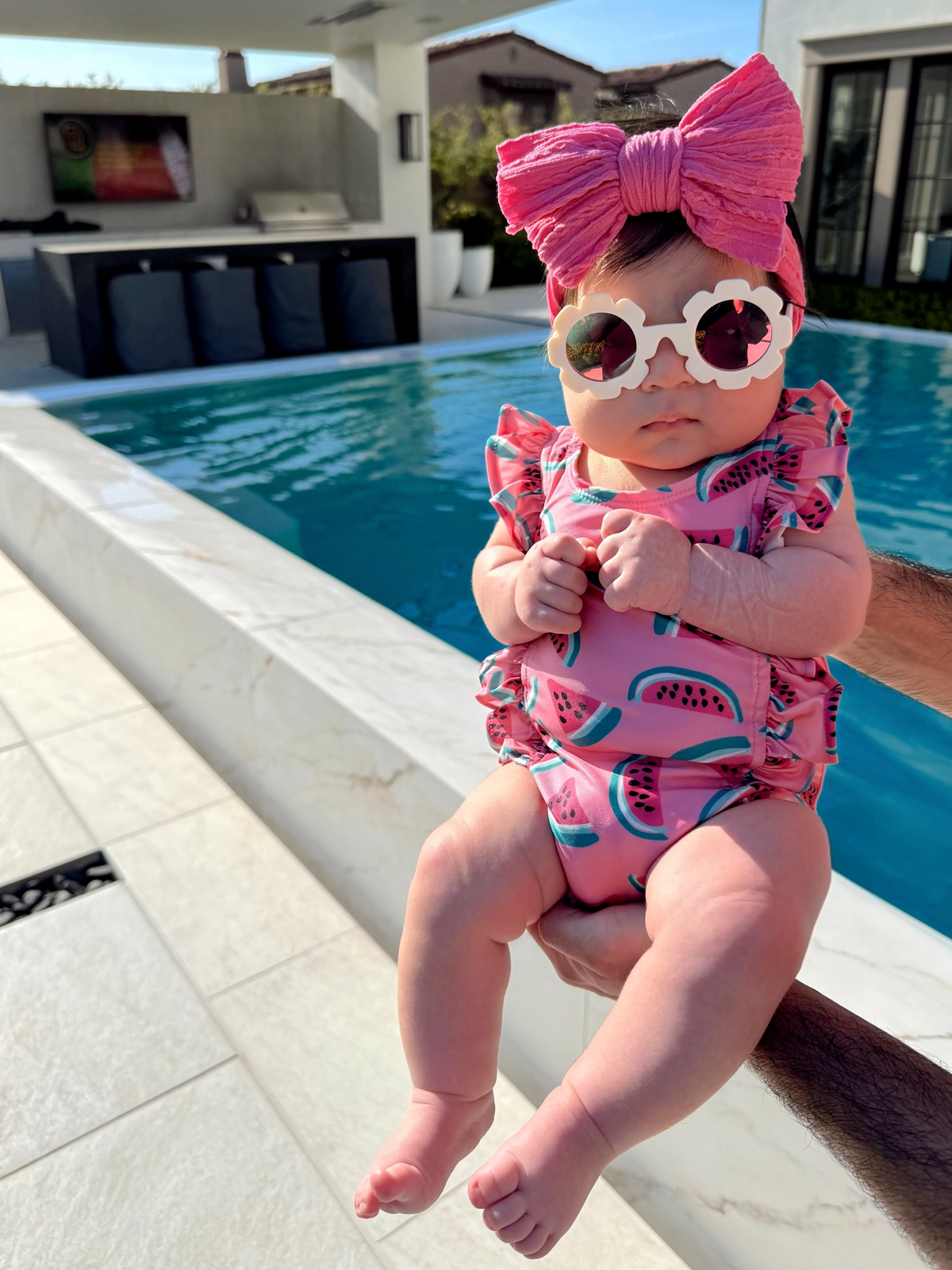 Baby wearing a pink watermelon swimsuit and large pink bow, held by a person beside a pool on a sunny day.
