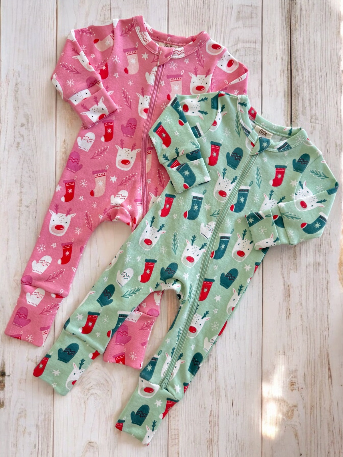 Two colorful Christmas-themed baby onesies, one pink and one green, displayed on a wooden surface.