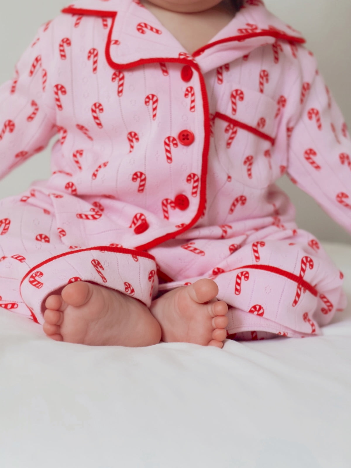 Child sitting on a bed wearing festive pink candy cane pajamas, with their feet visible on a white blanket.