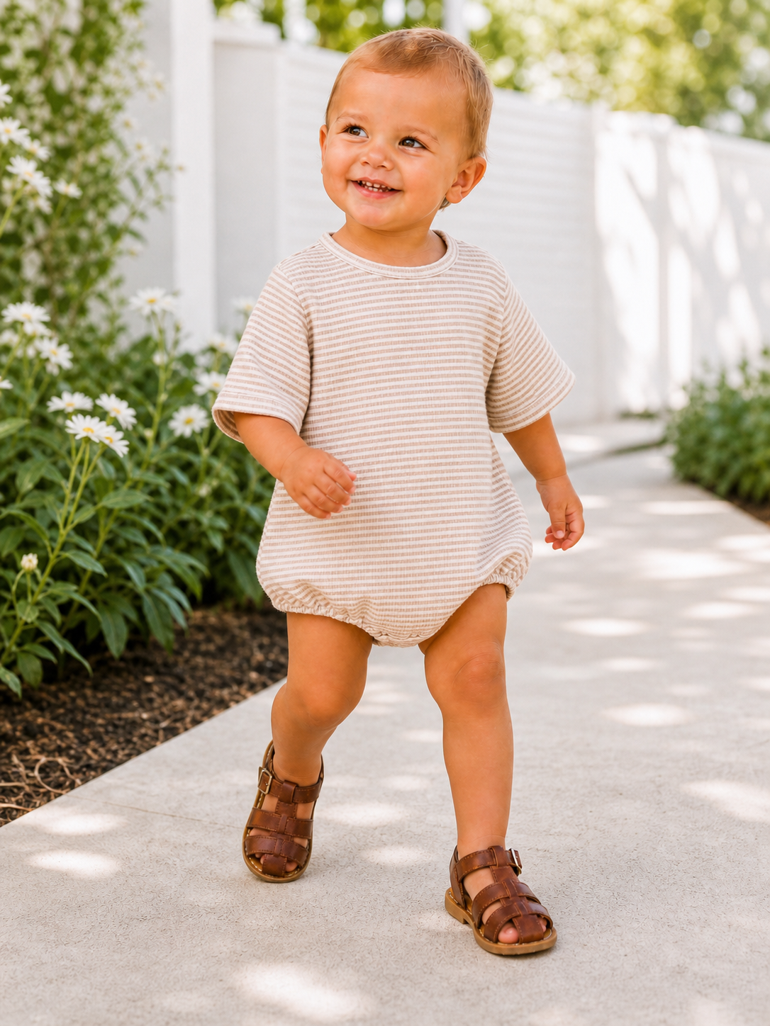 Smiling toddler in a striped outfit walks along a path with flowers and greenery in a bright, sunny setting.