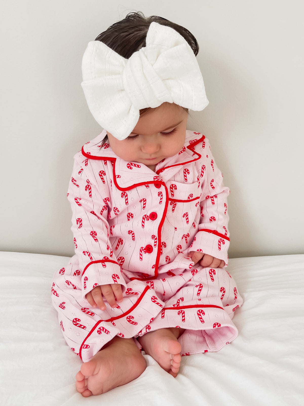 Baby sitting on a bed, wearing a candy cane patterned pink outfit and a large white bow headband.