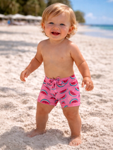 Smiling toddler in pink watermelon-patterned swim trunks playing in soft beach sand.