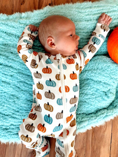 Baby lying on a turquoise blanket, wearing a pumpkin-patterned onesie, with a pumpkin toy nearby.