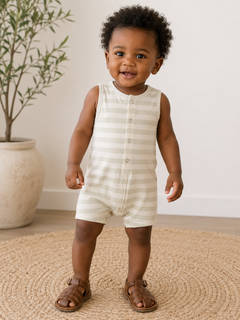 Smiling toddler in striped romper stands on a jute rug, with a potted plant in the background.