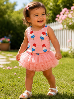Smiling toddler in a pink ice cream-themed top and tutu, playing on grass with blooming flowers in the background.
