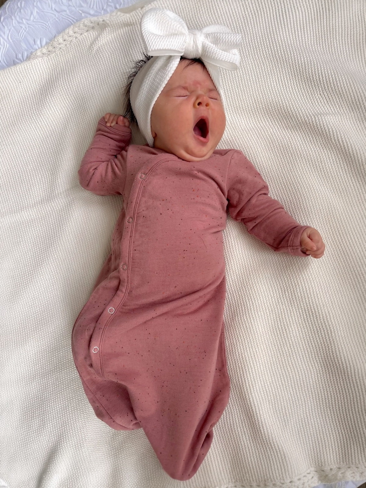 Baby yawning while lying on a cozy blanket, wearing a pink onesie and a large white bow headband.