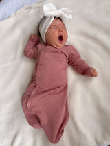 Baby yawning while lying on a cozy blanket, wearing a pink onesie and a large white bow headband.