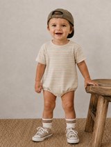 Smiling toddler in striped outfit and cap, standing beside a wooden stool on a textured surface.