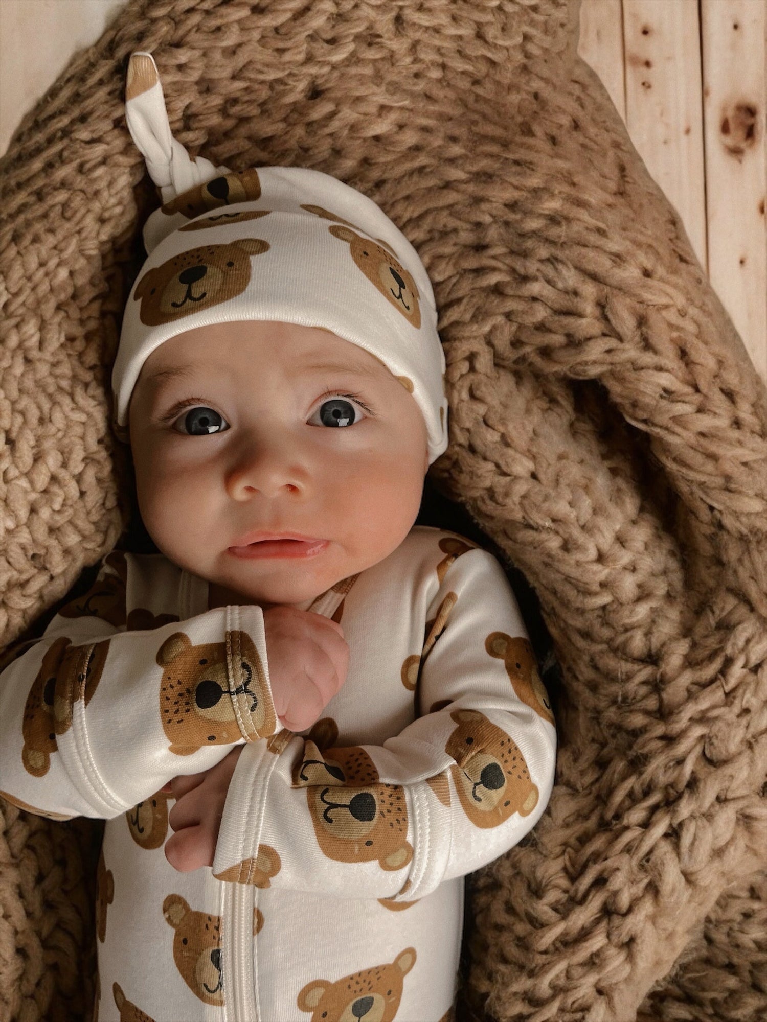 Baby in bear-patterned outfit and hat, lying on a cozy knitted blanket, with big expressive eyes.
