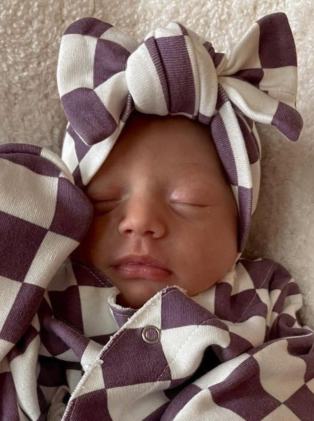 Infant sleeping peacefully in a checkered outfit and bow, featuring purple and white colors on a soft background.