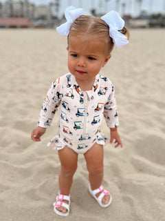 Toddler in a beach outfit with golf cart pattern, wearing a big bow, standing on sandy beach.