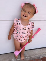 Smiling toddler in a pink golf cart patterned swimsuit, holding a pink golf club, with matching hair bows.