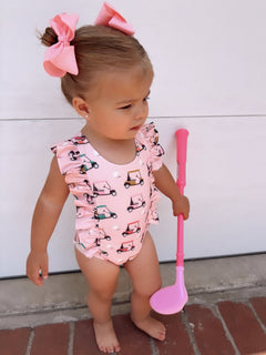 Toddler with pink bow and golf-themed swimsuit, holding a pink golf club, standing on brick pavement.