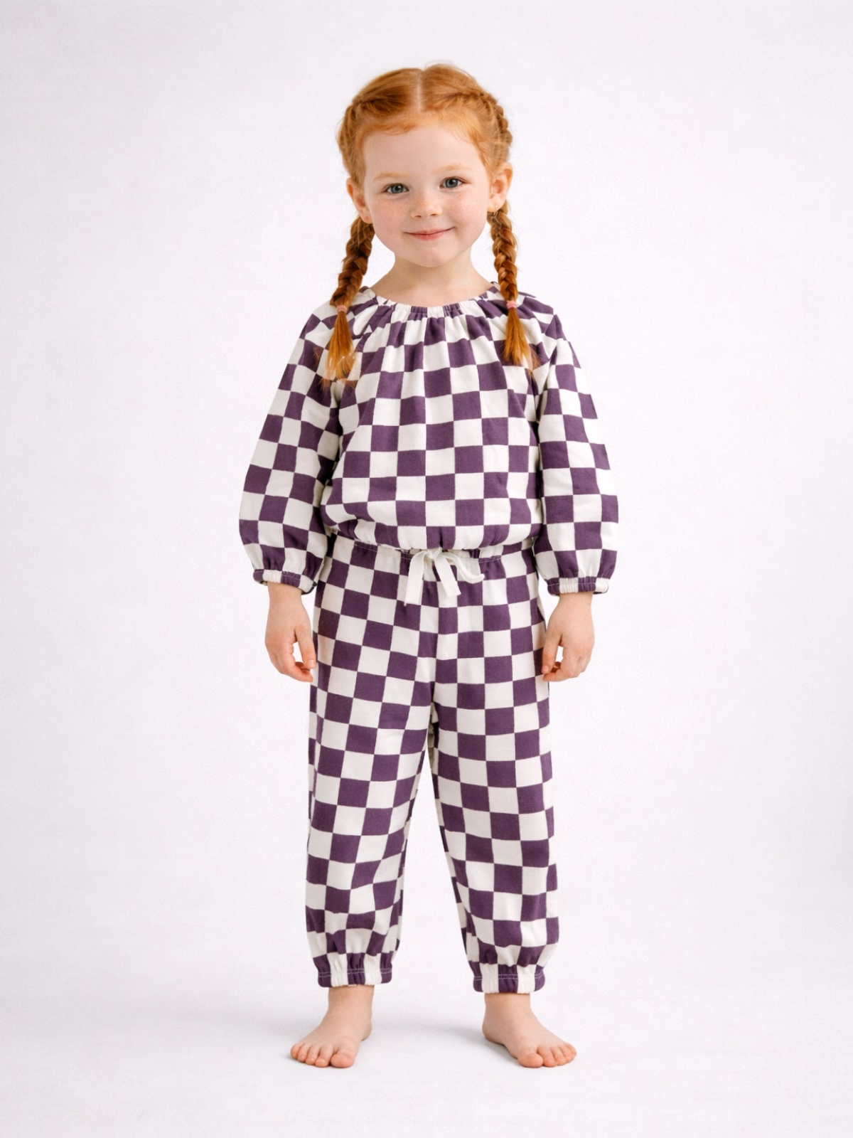 Smiling child with braided hair wearing a purple and white checkered outfit, standing barefoot on a light background.