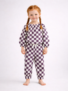 Smiling child with braided hair wearing a purple and white checkered outfit, standing barefoot on a light background.