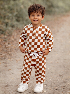 Smiling child in brown and white checkered outfit standing on a gravel path, wearing white sneakers.